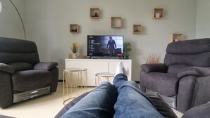 Un salon avec deux fauteuils inclinables gris, un meuble de rangement blanc avec un téléviseur à écran plat, et une petite table sur laquelle est posée une plante.