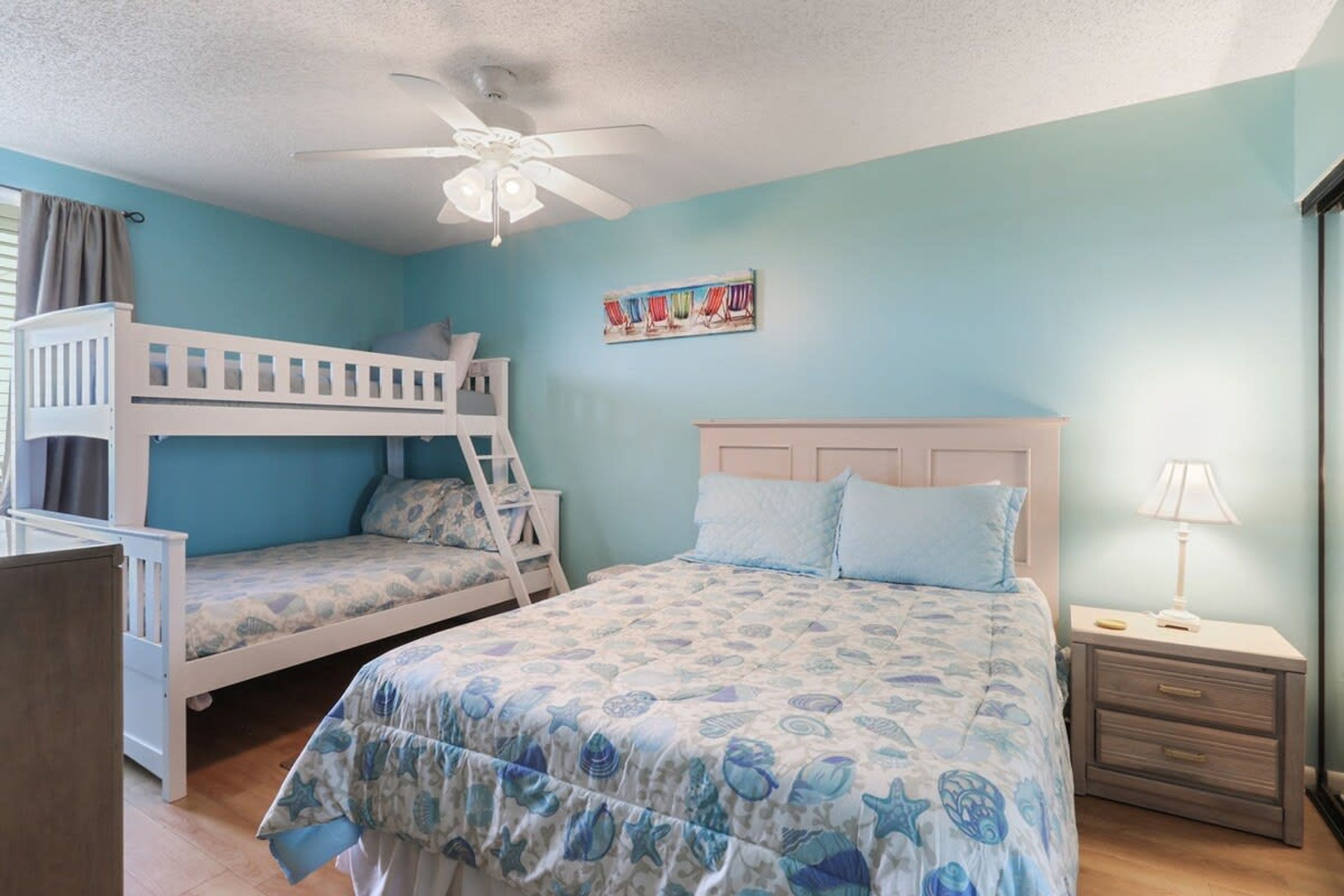 A bedroom with two bunk beds, a single bed, a ceiling fan, a nightstand with a lamp, and a wall painting.