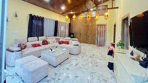 A modern living room with a sectional sofa, a coffee table, and a TV. The room has wooden ceiling beams and hanging lanterns. - GoBravo 37 - 2Bhk Farm with Pool & Garden Sec150 Noida (Noida)