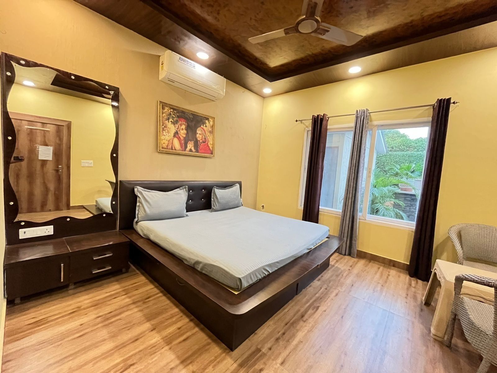 A bedroom with a bed, a dresser, a ceiling fan, an air conditioner, a window with curtains, and a painting on the wall.