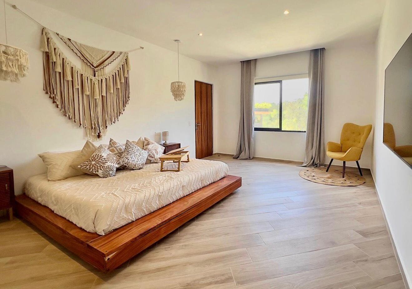 A modern bedroom with a bed, a yellow chair, a macrame wall hanging, and a large window.