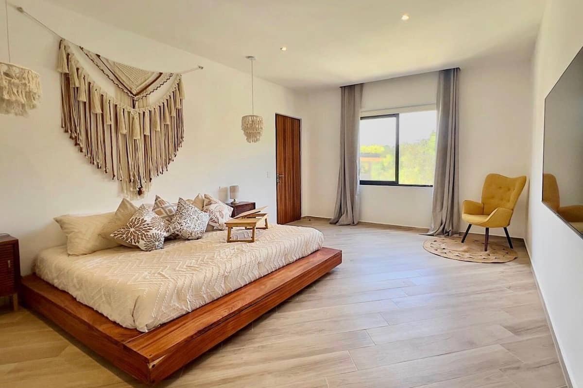 A modern bedroom with a bed, a yellow chair, a macrame wall hanging, and a large window.