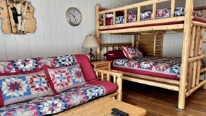 A bunk bed room with a wooden floor, a sofa with a floral blanket, a small table with a remote control, and a wall clock.