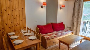 Habitació acollidora amb una taula de dinar de fusta preparada per a quatre, un sofà amb coixins vermells i dues làmpades penjades a la paret.