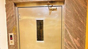 Puerta metálica de ascensor con un pequeño ventanal, panel de control con indicador rojo y pared con diseño de mármol.