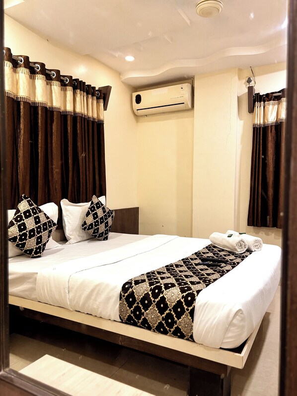 A neatly made bed with patterned pillows and a decorative throw, a wall-mounted air conditioner, and dark curtains. - Hotel Sai Balaji (Rahata)