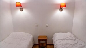 Una habitación pequeña y sencilla con dos camas individuales, una mesita de noche de madera y dos lámparas de pared.