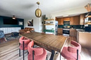 A modern kitchen with wooden cabinets, a central island, and a dining area with a long table and four pink chairs. - Siren's Oasis 3 Bedroom Pet Friendly St George Island (St George Island)