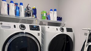 A laundry room with two front-loading washing machines, a shelf with various cleaning products, and a metal rack.