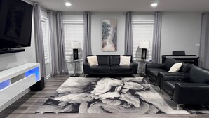 A modern living room with a black leather sofa, a flat-screen TV, a piano, and a large abstract rug.