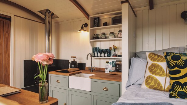 A cozy kitchen with a wooden countertop, a sink, and a stove. There are jars on a shelf above the stove and a vase with flowers on the countertop.