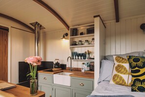 A cozy kitchen with a wooden countertop, a sink, and a stove. There are jars on a shelf above the stove and a vase with flowers on the countertop. - Luxury Shepherds Hut with Bath (Truro)