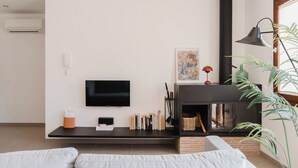A modern living room with a flat-screen TV, a fireplace, a bookshelf, and a large window with a plant. - La Figuera, house at Ultramort (Ultramort)