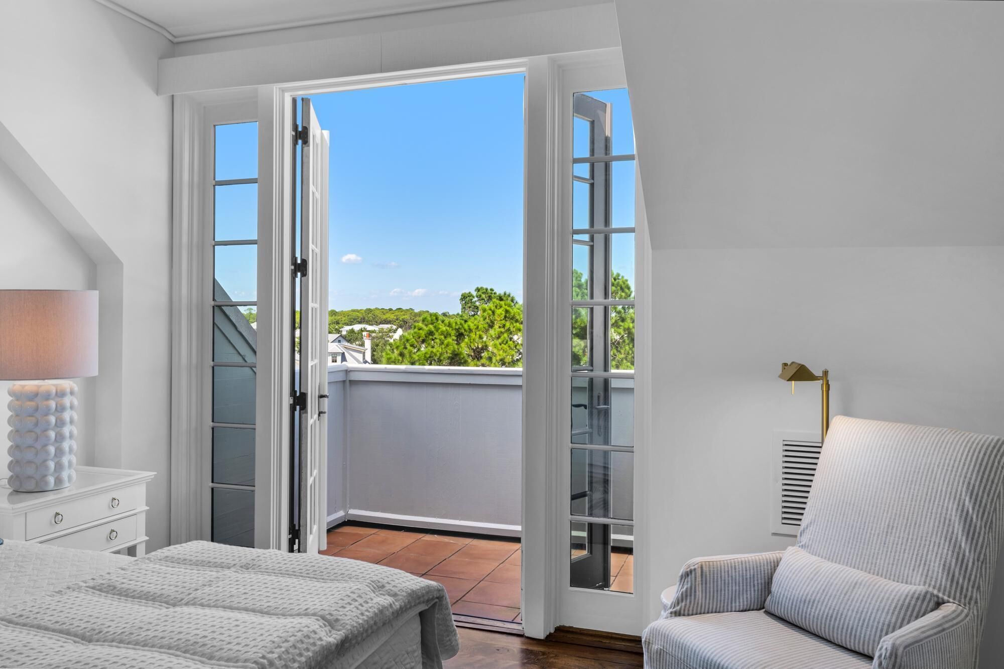 Camera da letto con un'ampia porta scorrevole in vetro che dà su un balcone. Si vede un letto con copriletto bianco, una lampada da comodino e una sedia con rivestimento a righe.