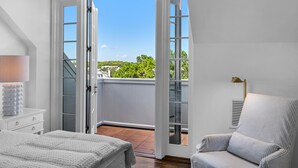 A bedroom with a large sliding glass door leading to a balcony. There is a bed with a white comforter, a bedside lamp, and a chair with striped upholstery.