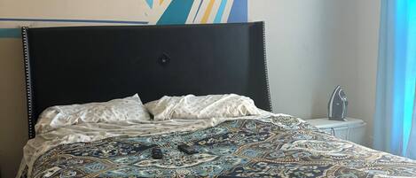 A neatly made bed with a patterned bedspread, a wall with geometric artwork, and a bedside table with a lamp.