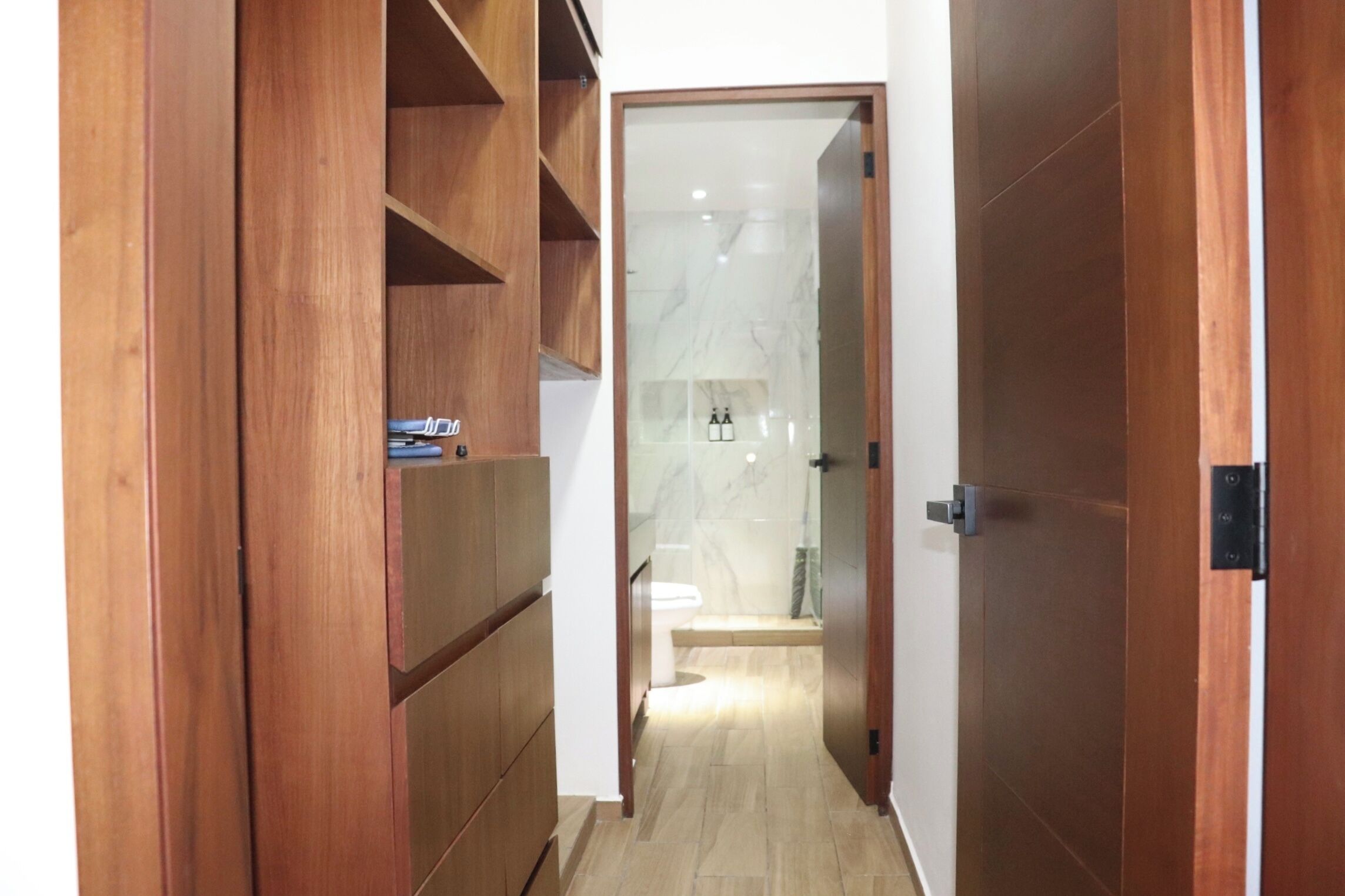 A hallway leading to a bathroom with a toilet and sink, wooden shelving, and a marble floor.