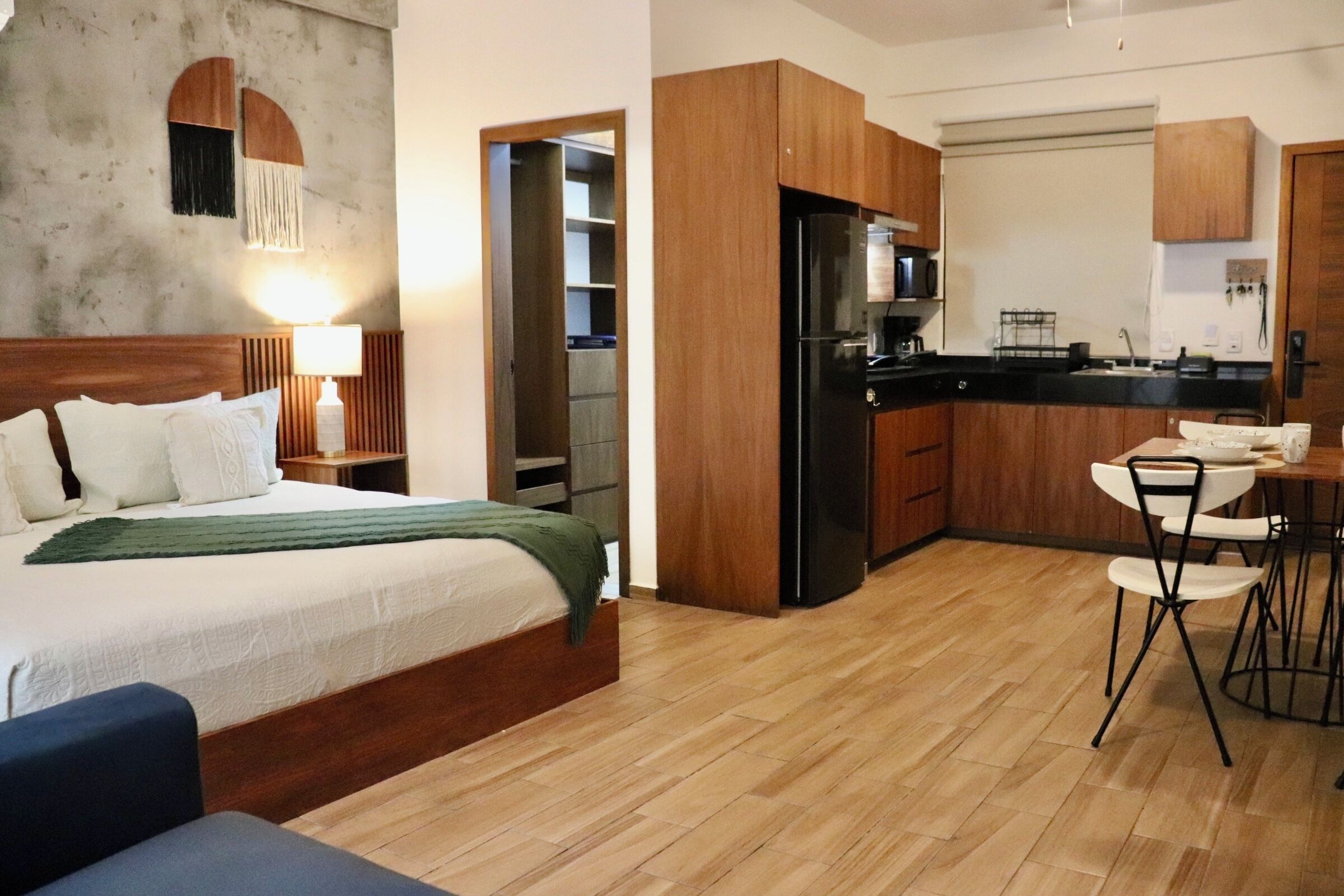 A hotel room with a bed, bedside table, lamp, and a view of the kitchen area.