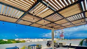 An outdoor patio area with a long table and chairs, a pergola overhead, and a view of the cityscape. - Sunset Kin Beach Apartments (Playa del Carmen)