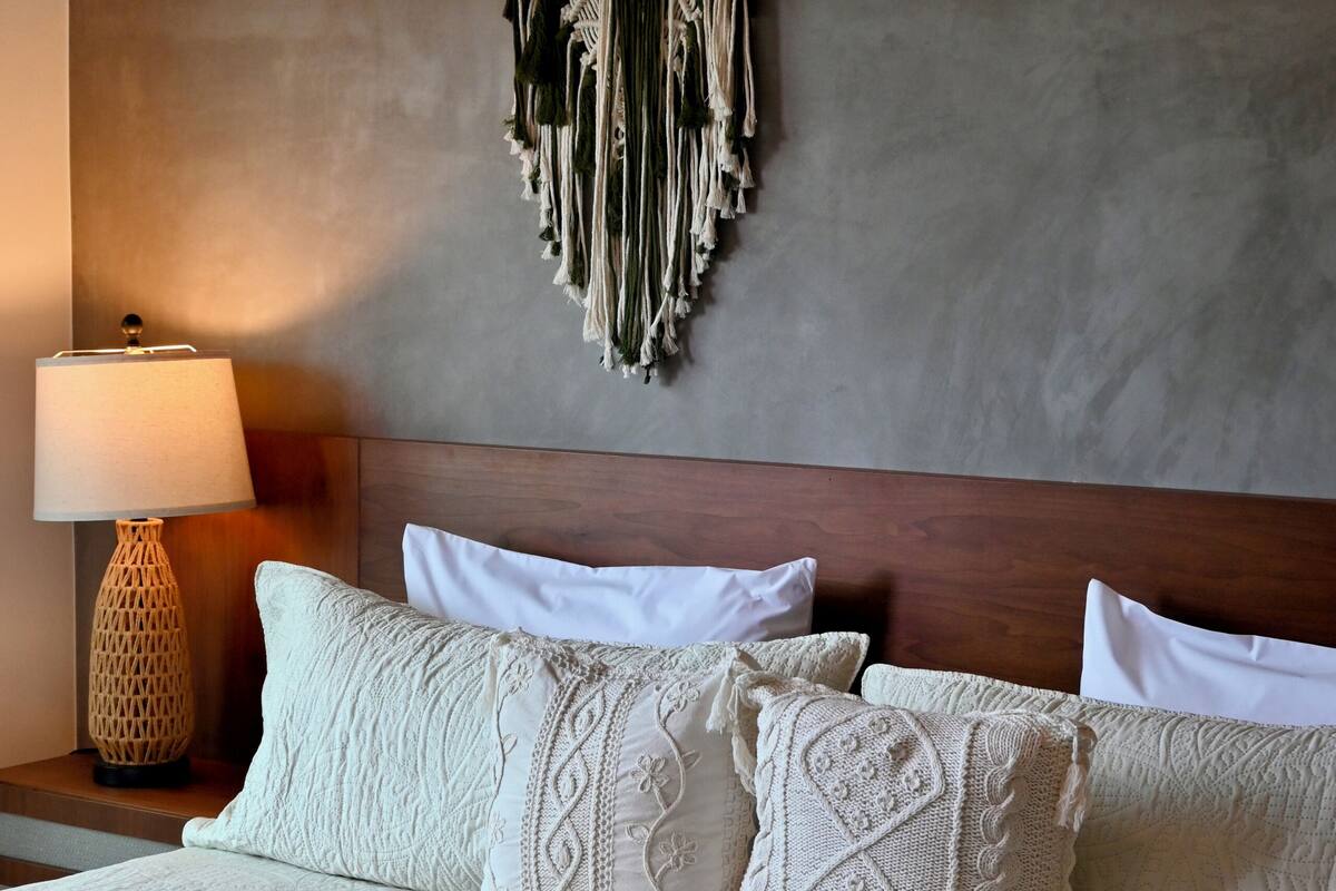 A neatly made bed with patterned pillows and a green throw blanket, a macrame wall hanging, and a lamp on the nightstand.
