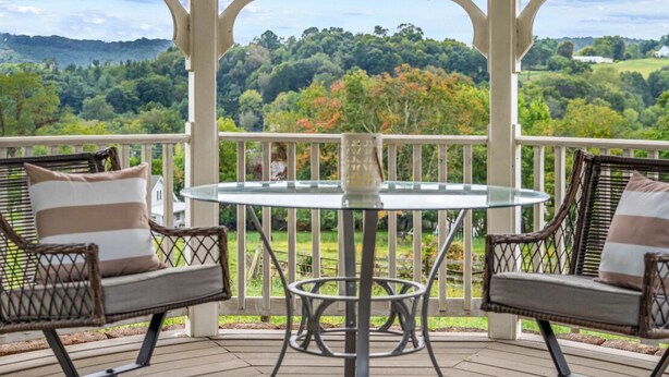 A covered outdoor seating area with wicker chairs and a glass table, overlooking a lush landscape.