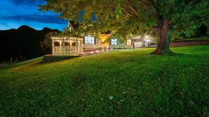 A well-lit gazebo-style structure with a lawn in front, surrounded by trees and a mountain backdrop.