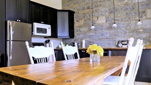 A kitchen with dark cabinets, a stainless steel refrigerator, a white microwave, and a stone wall. There is a wooden dining table with chairs and a vase of yellow flowers. - Luxury Limestone Suite is a Home Away from Home (KIngston)