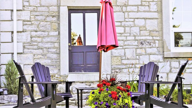 Un bâtiment en pierre avec une porte sombre, deux fauteuils Adirondack, une petite table et un parasol rose.