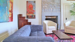 Un salon avec un canapé gris, deux fauteuils blancs, une cheminée et un grand tableau abstrait accroché au mur.