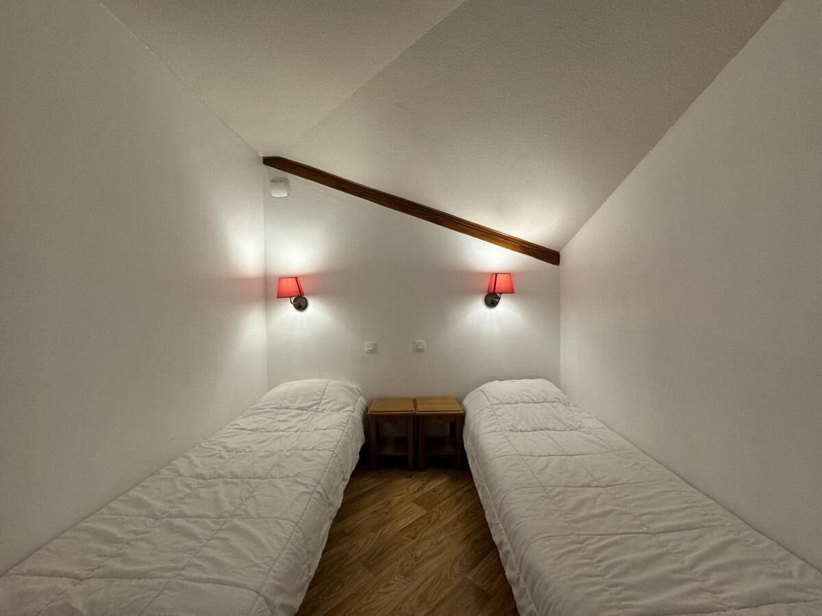 Una habitación pequeña y con poca luz, con dos camas individuales, una mesa de madera y dos lámparas fijadas a la pared.