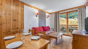 A wooden dining area with a table set for four, a sofa with red cushions, and a view of a forested landscape through large windows. - 40m from ski slopes, 3rd floor, fitness, swimming pool, sauna, tv, ski locker, 35m², Les Orres (Les Orres)