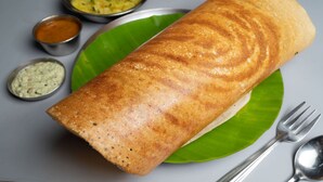 A rolled dosa on a green leaf with two small metal bowls of chutney and a spoon beside it. - La Pahi Palace (Madhubani)