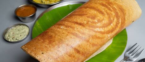 A rolled dosa on a green leaf with two small metal bowls of chutney and a spoon beside it.