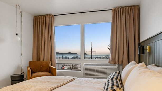 A hotel room with a bed, a chair, a window with a view of the sea, and an air conditioning unit. - Stylish Stay by Harbor | Walkable Hotspot (Juneau)