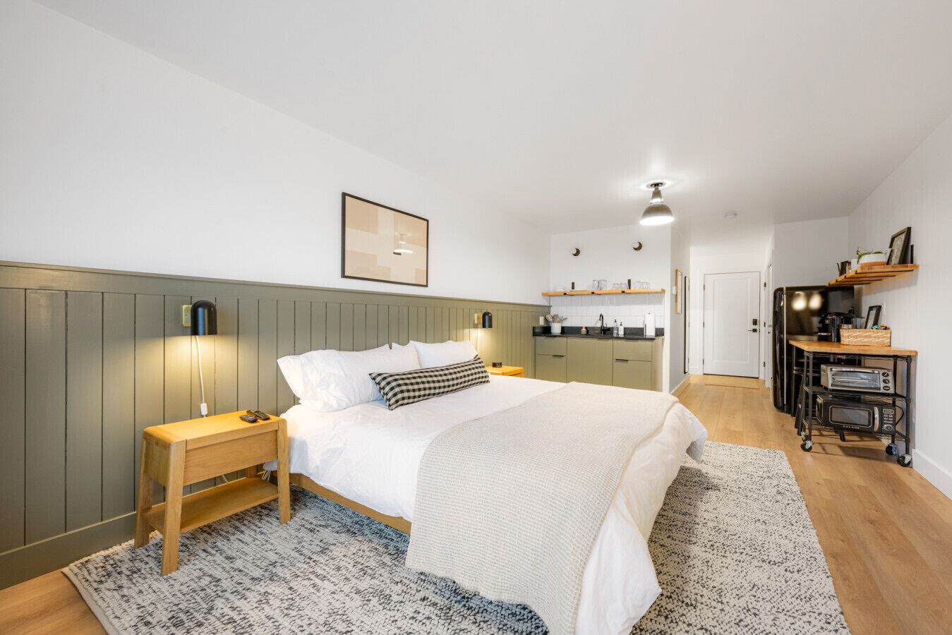 A modern bedroom with a bed, bedside tables, a nightstand, a desk, and a kitchenette area with a microwave and toaster.