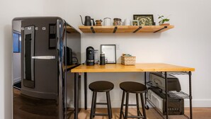 A compact kitchen with a refrigerator, a coffee machine, a wooden table, two stools, and a small shelf with kitchen items. - Stylish Stay by Harbor | Walkable Hotspot (Juneau)