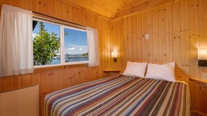 Ein Zimmer in einer Holzhütte mit einem Bett, einem Fenster mit Vorhängen und Blick auf einen Baum und Wasser.