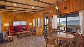 Ein gemütliches Blockhaus-Interieur mit Holzboden, einer roten Couch mit Kissen, einem Esstisch mit Stühlen und einem großen Fenster mit Blick auf das Wasser.