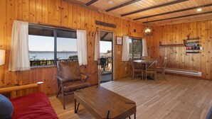 Eine Holzhütte mit Bett, Tisch und Stuhl. Ein Fenster bietet Blick auf das Wasser.