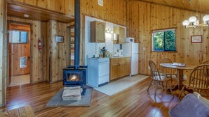 Interior de madera con estufa de leña, cocina con electrodomésticos blancos y comedor con sillas.