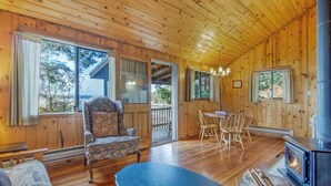 Interior acogedor de una cabaña con paredes y suelos de madera, zona de comedor, chimenea y ventanas amplias.