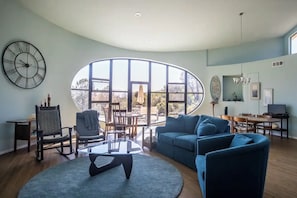 Ein geräumiges Wohnzimmer mit einem großen, ovalen Fenster, einem modernen Sofa, einem Glastisch und einem Essbereich mit einer Pendelleuchte.