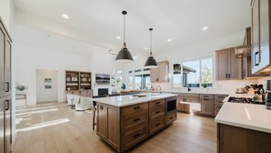 A modern kitchen with a central island, wooden cabinets, pendant lighting, and a large window providing natural light.