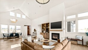 A spacious living room with a large brown sectional sofa, a fireplace, and a television mounted above it. There is a dining area with a table and chairs, and a kitchen area with wooden cabinets and a countertop.
