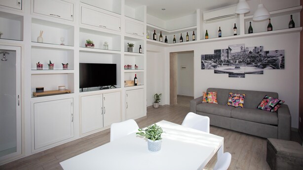 A modern living room with white cabinetry, a flat-screen TV, a grey sofa with colorful cushions, and a dining area with a white table and chairs. - La finestra sul Pincio, Appartamento a due passi dalla stazione e dal porto. (Civitavecchia)