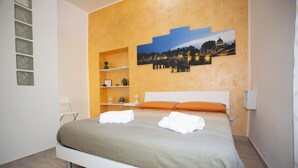 A modern bedroom with a bed, a nightstand, a shelf with decorative items, and a cityscape painting on the wall. - La finestra sul Pincio, Appartamento a due passi dalla stazione e dal porto. (Civitavecchia)