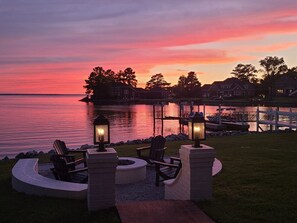 Une terrasse au bord de l’eau, dotée de fauteuils Adirondack et d  ’un foyer, offrant une vue sur un lac avec des maisons en arrière-plan.