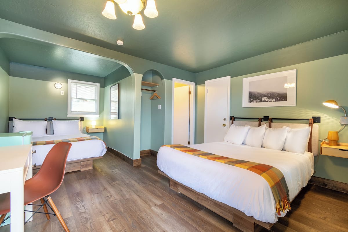 A room with two beds, a desk, and a chair. There is a window, a door, and a picture on the wall.