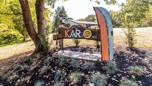 A wooden sign with the text "KAR" and a logo, surrounded by trees and a leaf-covered ground.