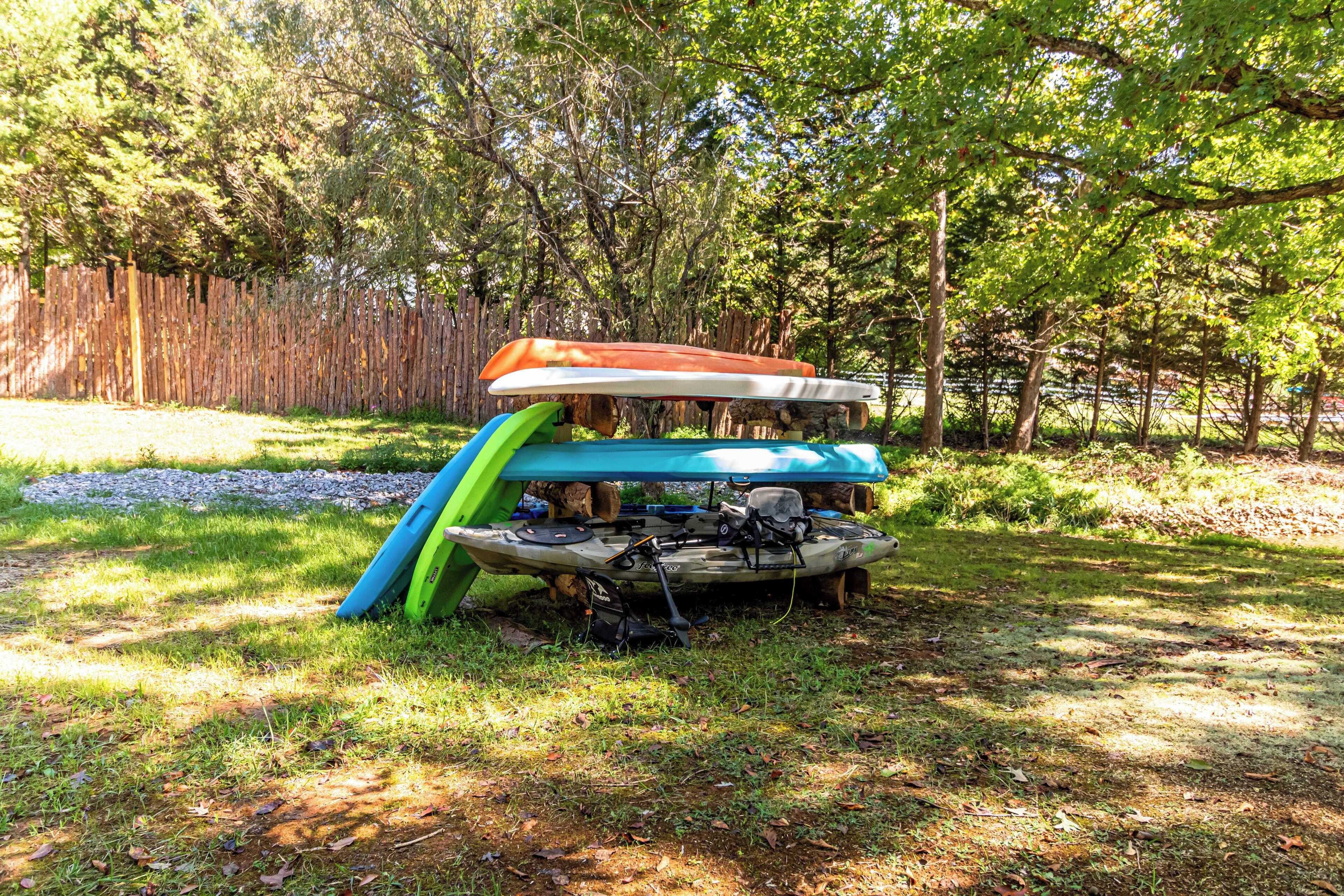 A stack of kayaks and a paddleboard in a grassy area with trees in the background.